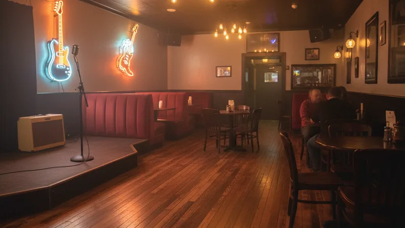 Warm interior of a small Nashville-style honky-tonk bar with neon signs glowing softly, a small stage with a microphone stand and a guitar amplifier, wooden floors and warm hanging Edison bulb lights, candid documentary photography, atmospheric warm amber tones, country music venue aesthetic